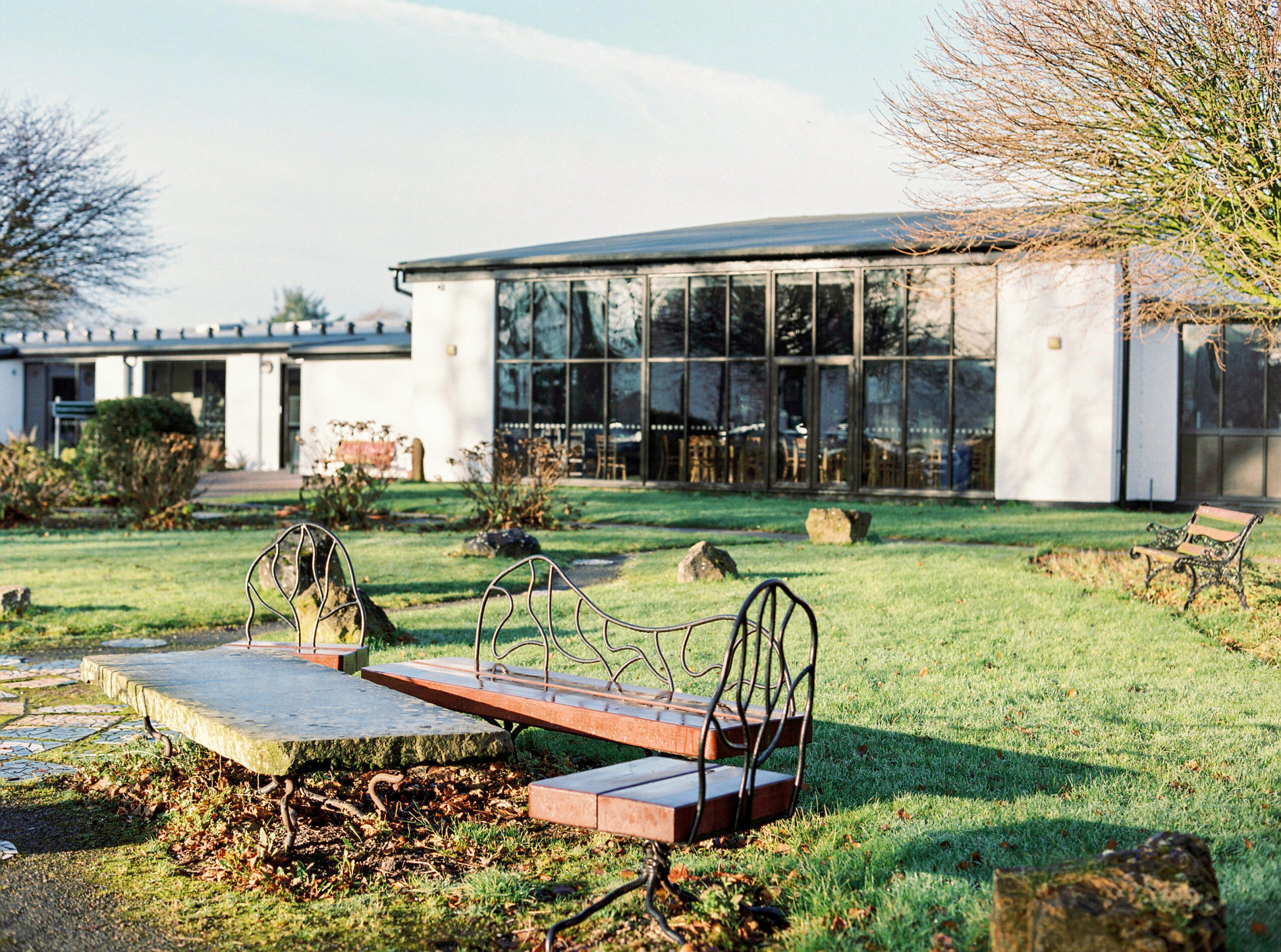 A white building with a large glass window and flat room. In front of the building is some grass and a seating area